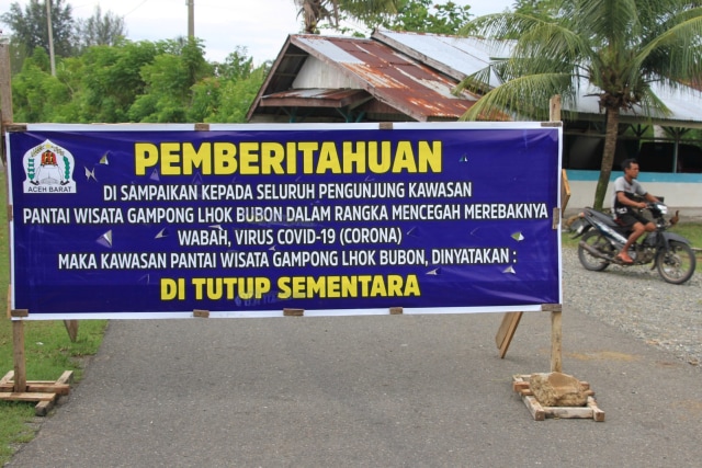 Seorang pengendara melintas di samping spanduk informasi penutupan pantai wisata di Desa Lhok Bubon, Kecamatan Samatiga, Aceh Barat, Aceh, Minggu (5/4). Foto: ANTARA FOTO/Syifa Yulinnas