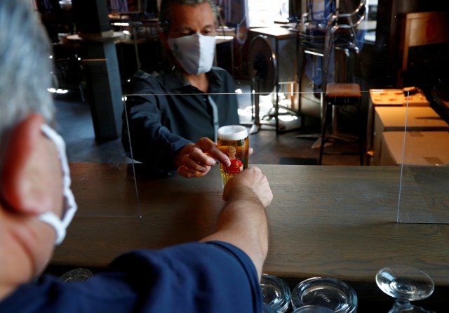 Pemilik bar Le Louvre memegang segelas bir saat ia menjajal layar plexiglass pembatas di konter barnya di Brussels, Belgia. Foto: REUTERS/Francois Lenoir