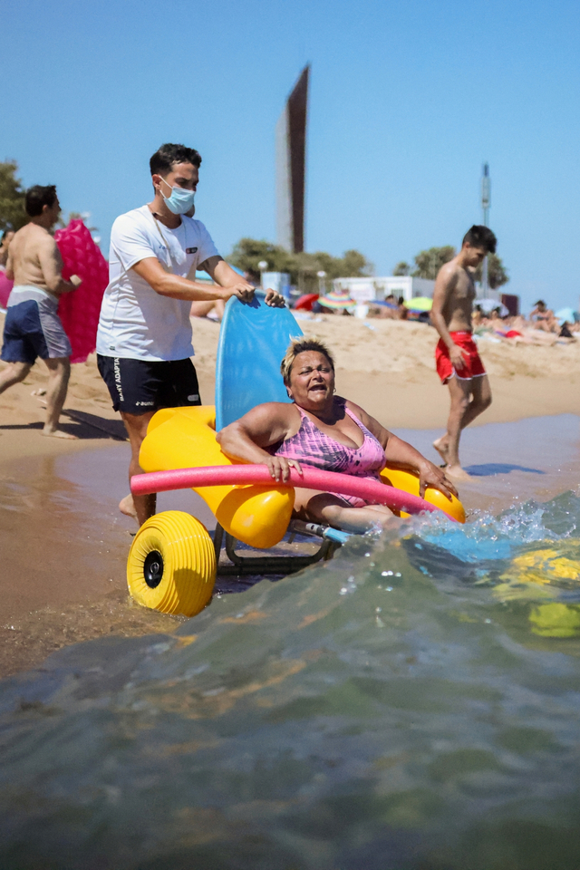 Seorang penyandang disabilitas duduk di kursi amfibi saat dibantu penjaga pantai di Pantai Nova Icaria Barcelona, Spanyol. Foto: Nacho Doce/REUTERS