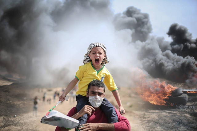 Foto anak laki-laki digendong seorang laki-laki dewasa dengan latar belakang api ledakan bom. Foto: Diambil dari website PIXABAY (https://pixabay.com/id/images/search/palestina/).