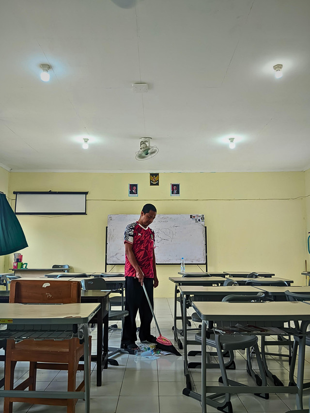 Petugas kebersihan sekolah sedang membersihkan salah satu kelas yang kotor.