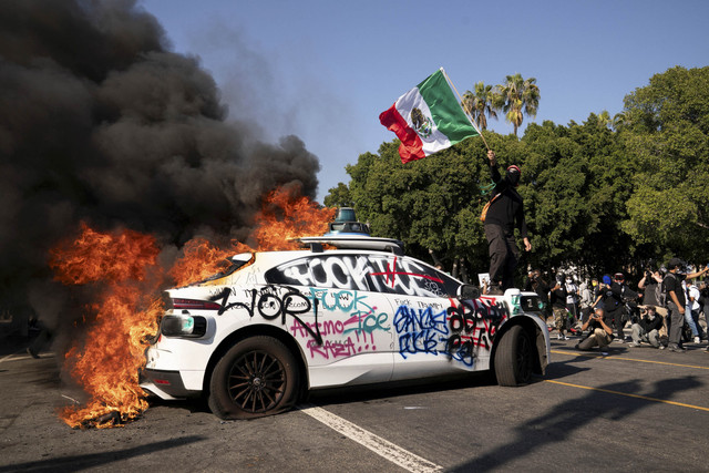 Seseorang melambaikan bendera Meksiko di atas kendaraan yang terbakar selama protes terhadap razia imigrasi federal di pusat kota Los Angeles, California, AS, Minggu (8/6/2025). Foto: David Ryder/REUTERS