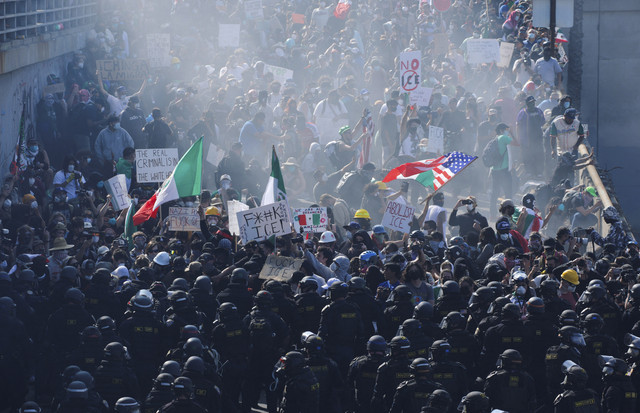 Para demonstran melambaikan bendera dan menunjukkan tanda-tanda saat mereka berhadapan dengan polisi di tengah asap selama protes terhadap razia imigrasi federal di pusat kota Los Angeles, California, AS, Minggu (8/6/2025). Foto: Mike Blake/REUTERS