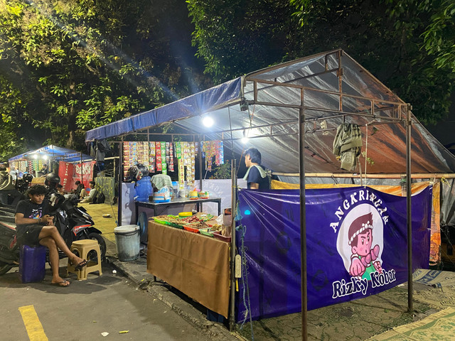 Mas Rizky berdiri di balik meja angkringan, menunggu pelanggan yang datang