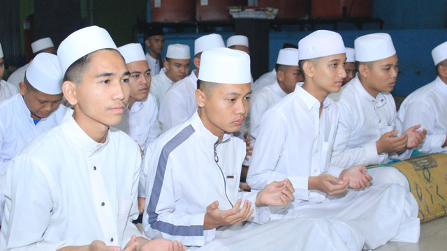 Gambar ini menampilkan sekelompok pemuda, kemungkinan besar santri, yang mengenakan pakaian muslim serba putih dengan peci. Mereka terlihat sedang duduk di lantai dengan posisi tangan menengadah, mengisyaratkan sedang berdoa atau berzikir. Ekspresi wajah mereka tampak fokus dan khusyuk. Latar belakang menunjukkan dinding sederhana, mengindikasikan bahwa mereka berada di dalam ruangan. (Foto ini diambil dari galeri pribadi)