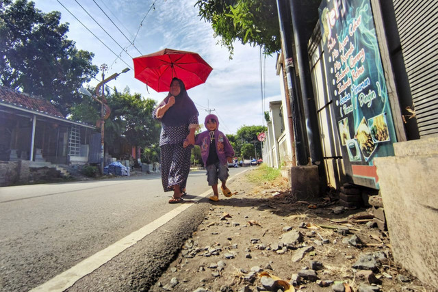 Kartini dan anaknya berjalan menggunakan payung saat cuaca panas di Jalan D.I. Panjaitan, Kabupaten Indramayu, Selasa (21/10/2025). Foto: Dok. kumparan