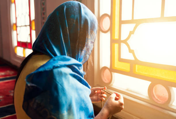 Wanita muslim berhijab dan berhijab berdoa dengan tangan di udara sambil memegang tasbih di masjid. Foto by Istock