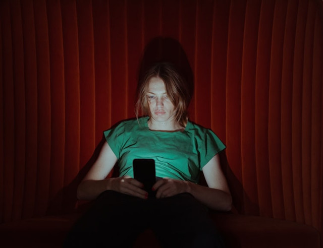 Seorang perempuan muda larut dalam layar ponselnya di kegelapan, menggambarkan bagaimana media sosial menjadi sumber utama informasi kesehatan mental bagi generasi muda. (Foto: Unsplash)