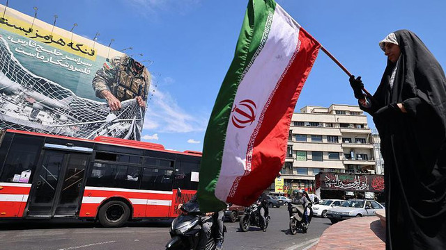 Seorang perempuan mengibarkan bendera nasional Iran di depan papan reklame raksasa bertuliskan 'Selat Hormuz tetap ditutup' di Lapangan Revolusi, Teheran. Minggu (12/04).