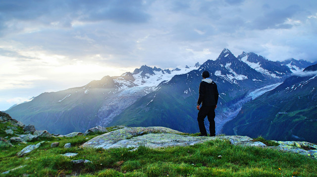 Seorang pria berdiri di puncak gunung menghadap hamparan pegunungan, menggambarkan perubahan standar hidup dan ekspektasi sosial. Sumber: Nicolas Cool/Unsplash.