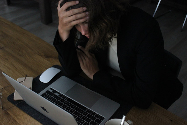 Seorang perempuan tampak kelelahan sambil memegang ponsel di depan laptop — menggambarkan momen yang sangat familiar: tahu harus berhenti scroll, tapi tangan bergerak sendiri. Kondisi ini bukan soal lemahnya kemauan, melainkan bagaimana sistem reward di otak bekerja secara otomatis di luar kendali kesadaran penuh. (Foto: Nama Fotografer via Unsplash)