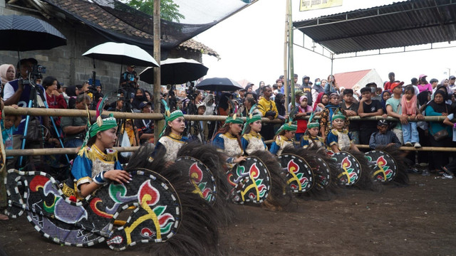 Sejumlah penari jatilan bersiap di arena pertunjukan dengan properti kuda lumping bercorak warna-warni, disaksikan oleh kerumunan penonton yang memadati pagar pembatas di bawah deretan payung dan tenda.(Foto: Dokumentasi Pribadi)