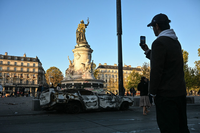 Seorang pejalan kaki mengambil foto kendaraan yang terbakar dan patung 'Monumen a la Republique' yang memperlihatkan Rompi Kuning sehari setelah protes nasional menentang "tingginya biaya hidup", di Paris, Minggu (18/10/2026). Foto: MATTHIEU RONDEL/AFP