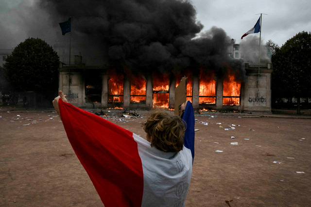 Pengunjuk rasa berselimut bendera Prancis berdiri di depan kantor pariwisata Lyon yang terbakar saat demonstrasi menolak tingginya biaya hidup di Lyon, Prancis, Sabtu (17/10/2026). Foto: Jeff Pachoud/AFP