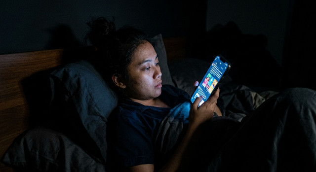 seseorang menatap layar ponsel di dalam kamar yang gelap gulita pada malam hari, merepresentasikan kebiasaan overstimulated dan doomscrolling. Sumber : Gemini AI