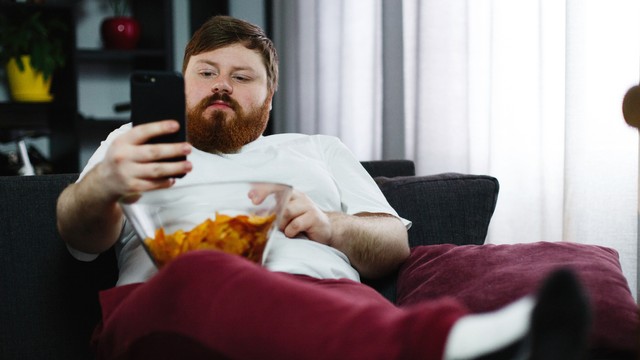 Pandemi memicu terjadinya obesitas. Foto: https://www.freepik.com/free-photo/pretty-fat-man-smiles-checking-his-smartphone-while-he-sits-sofa-eats_2446949.htm#page=1&query=obesity&position=7&from_view=search