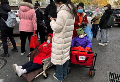 Orang-orang berdiri di samping anak-anak yang duduk di kereta kemah saat mereka menunggu perjalanan di luar rumah sakit anak-anak di Beijing, China, Senin (27/11/2023). Foto: Tingshu Wang/REUTERS