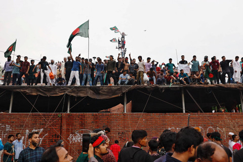 Seorang pria yang memegang bendera Bangladesh berdiri di depan sebuah kendaraan yang dibakar di Ganabhaban, kediaman Perdana Menteri, setelah pengunduran diri PM Sheikh Hasina di Dhaka, Bangladesh, 5 Agustus 2024. Foto: Mohammad Ponir Hossain/Reuters