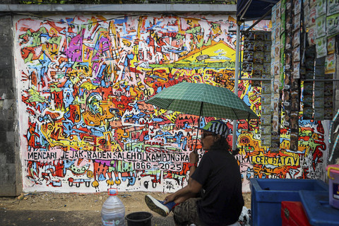 Seniman duduk di depan karya mural di kawasan Kalipasir, Cikini, Jakarta, Kamis (24/7/2025). Foto: Iqbal Firdaus/kumparan