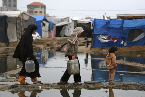 Para perempuan Palestina yang mengungsi menggunakan jalan kering untuk mengangkut air ke tenda mereka, menyusul hujan deras di utara Deir el-Balah di Jalur Gaza tengah (24/11/2025). Foto: Bashar Taleb/AFP