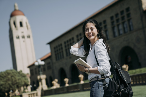 Seorang mahasiswi Asia belajar di luar negeri, membawa ransel dan buku pelajaran, berdiri di luar kampus universitas di hari cerah di Amerika. Foto: Shutterstock
