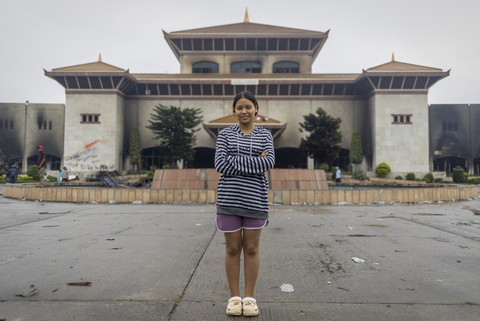 Seorang pekerja pabrik garmen, Srijana Bhujel (19) berpose dengan latar belakang gedung Parlemen yang terbakar di vandalisme saat mengikuti aksi menentang korupsi di Kathmandu, Nepal. Foto: Navesh Chitrakar/Reuters