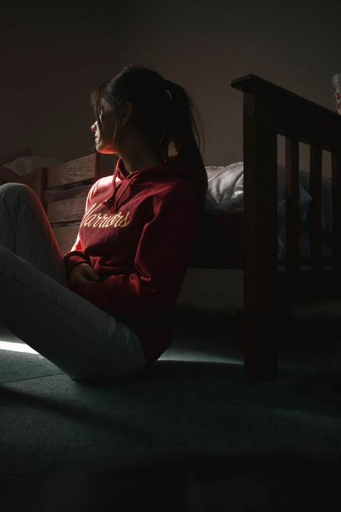 Seorang remaja perempuan duduk di lantai tamaram, menatap cahaya dengan ekspresi sendu. Sumber: Pexels, Sofia Alejandra.