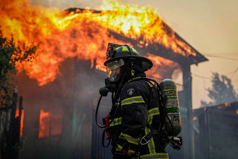 Petugas pemadam kebakaran berdiri di depan bangunan yang terbakar saat api dan asap mengepul dari kebakaran hutan di wilayah Biobio, tempat menurut media lokal, beberapa kebakaran hutan memicu evakuasi darurat, di Concepcion, Chili, (18/1/2026). Foto: REUTERS/Juan Gonzalez 