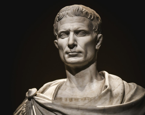 Patung buste Julius Caesar dengan latar belakang gelap, menunjukkan detail wajah dan jubah Romawi. Foto: Sam Szuchan via Unsplash