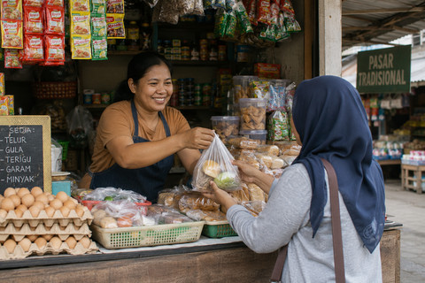 Ilustrasi : Foto pelaku usaha kecil di kios atau warung yang sedang melayani pembeli, dengan suasana pasar tradisional (sumber : chatgpt.com)