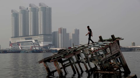 Seorang pemuda berjalan di atas puing-puing perahu kayu dengan latar gedung di Jakarta Utara, Indonesia. Foto: REUTERS/Beawiharta