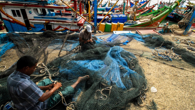 Nelayan memperbaiki jaring di dekat sejumlah kapal motor yang tidak digunakan untuk melaut di Tempat Pendaratan Ikan (TPI) Pusong, Lhokseumawe, Aceh, Selasa (25/8/2018). (Foto: ANTARA FOTO/Rahmad)