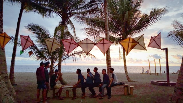 Para pemuda berfoto di sebuah kursi kayu yang memanjang. Dengan lanskap langit merah juga pepohonan kelapa. Foto: Rajif Duchlun/cermat