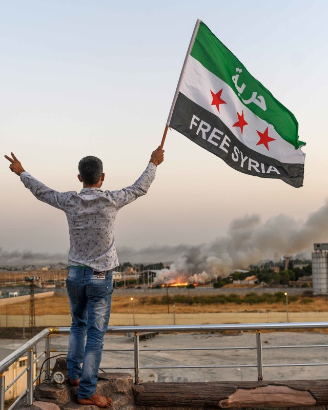 Seorang pria mengibarkan bendera oposisi Suriah bertuliskan "Bebaskan Suriah" (13/10/2019) di Akcakale ketika asap naik di latar belakang dari kota Tal Talyyad. Foto: AFP/Nazeer Al-khatib