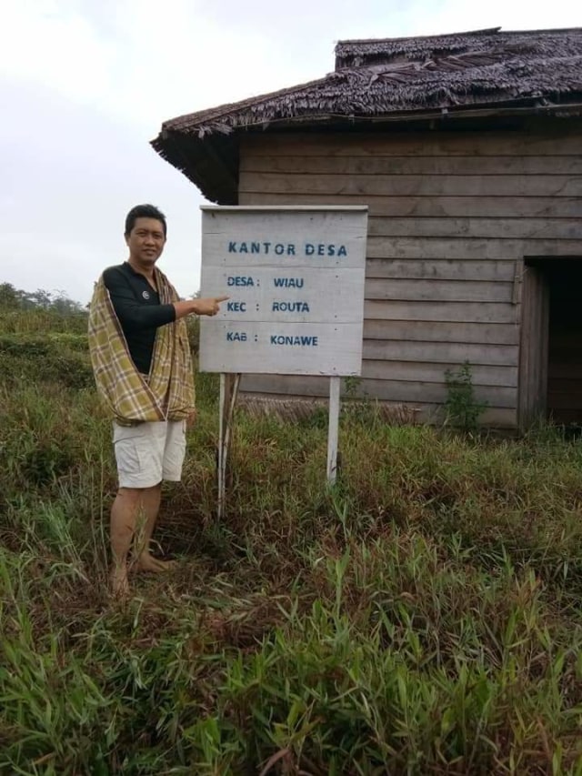 Salah seorang pengunjung menunjukkan papan nama kantor desa yang diduga siluman yang berada di Kabupaten Konawe, Sulawesi Tenggara. Di papan mana itu tertulis jelas nama desa Wiau, Kecamatan Routa. Foto: Istimewa.