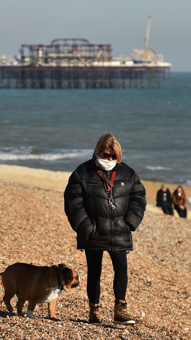 Seorang wanita menggunakan masker berjalan bersama anjing di pantai di Brighton, Inggris selatan. Foto: AFP/ Glyn KIRK