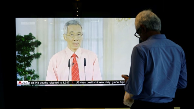 Seorang pria berdiri di depan layar yang memperlihatkan siaran Perdana Menteri Singapura Lee Hsien Loong menyikapi wabah virus corona. Foto: REUTERS/Edgar Su