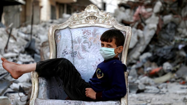 Seorang anak duduk di sofa di tengah reruntuhan bangunan, di kota Ariha di pedesaan selatan provinsi Idlib, Suriah, 11 April 2020. Foto: AFP/AAREF WATAD