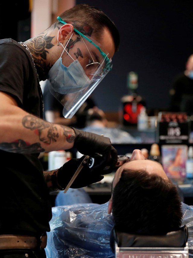 Seorang tukang cukur mengenakan masker dan pelindung wajah saat memotong janggut pelanggan di Barber Shop di Paris. Foto: REUTERS / Benoit Tessier