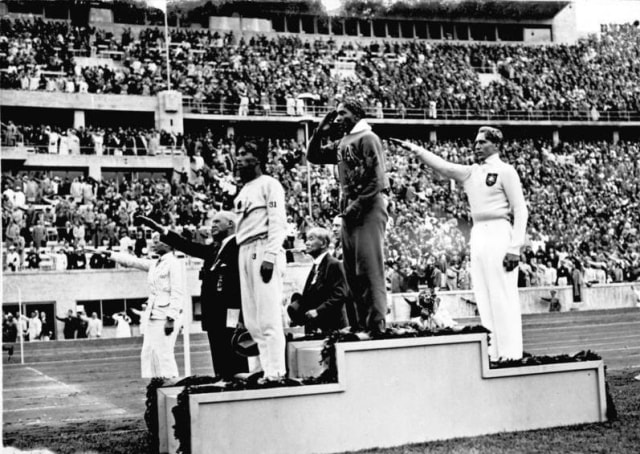 Jesse Owens (di atas podium-tengah) usai memenangkan  lompat jauh di Olimpiade Berlin 1936. Ia mengalahkan atlet Jerman, Luz Long (kanan), yang juga sahabatnya. Naoto Tajima dari Jepang di posisi ketiga. Foto: Dok. Wikimedia Commons.   