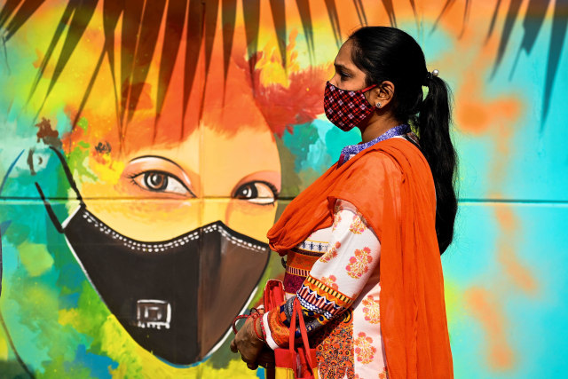 Seorang wanita mengenakan masker berdiri di depan mural bergambar ajakan menggunakan masker di New Delhi, India. Foto: Sajjad Hussain/AFP