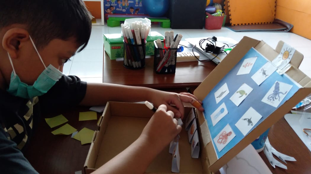 Siswa sedang membuat diorama dengan arahan guru pendamping sebagai bentuk assessmen dalam memahami lingkungan sekitar dengan tema laut saat kegiatan stimulasi dengan protokol kesehatan yang ketat.