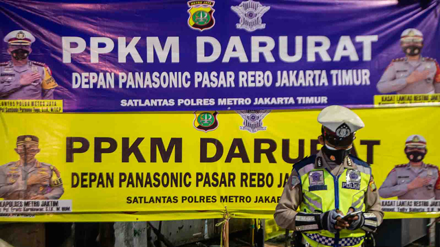 Seorang Polisi berdiri di depan baliho pengumuman pelaksanaan Pemberlakuan Pembatasan Kegiatan Masyarakat (PPKM) Darurat di Jalan Raya Bogor, Jakarta. Foto: M RISYAL HIDAYAT/ANTARA FOTO
