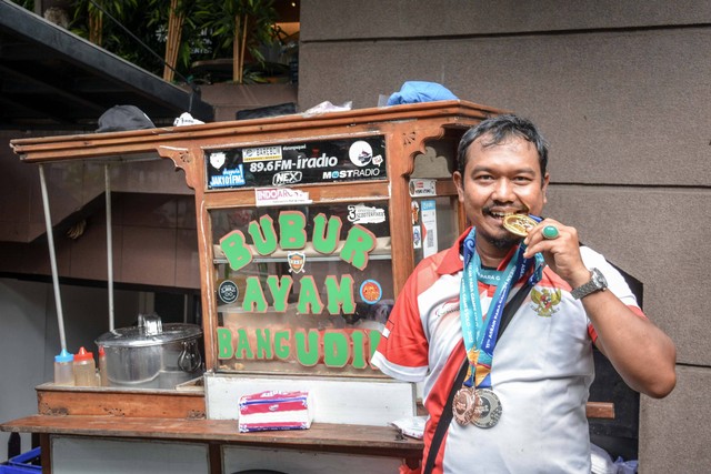 Kholidin alias Bang Udin, atlet parapanahan yang raih medali ASEAN Para Games 2022, berpose di tempatnya berjualan bubur di parkiran Wisma Geha, Jakarta Pusat, pada Agustus 2022. Foto: Alif Zaky Assidiqi/kumparan
