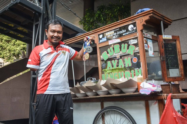 Kholidin alias Bang Udin, atlet parapanahan yang raih medali ASEAN Para Games 2022, berpose di tempatnya berjualan bubur di parkiran Wisma Geha, Jakarta Pusat, pada Agustus 2022. Foto: Alif Zaky Assidiqi/kumparan