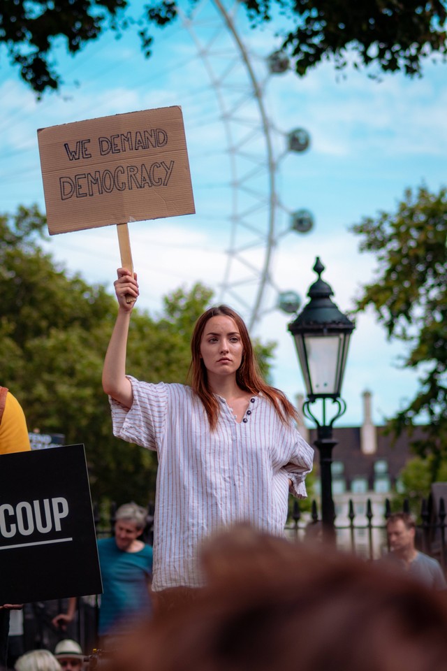 Seorang perempuan memegang papan bertuliskan "kami menuntut demokrasi" (Foto oleh Fred Moon/Unsplash)