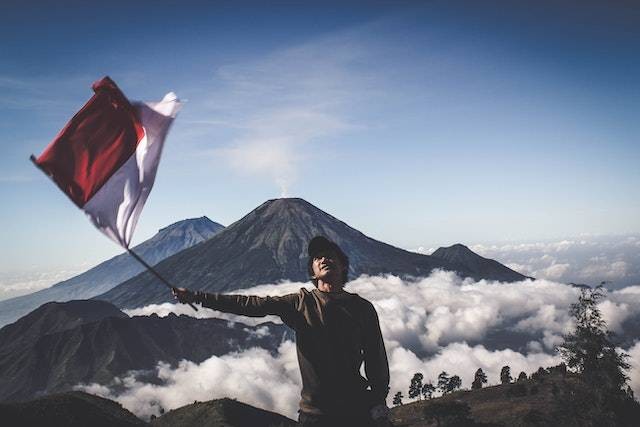 Sumber: https://www.pexels.com/id-id/foto/pria-mengenakan-sweater-leher-kru-hitam-memegang-bendera-putih-dan-merah-berdiri-dekat-gunung-di-bawah-langit-biru-dan-putih-1009949/