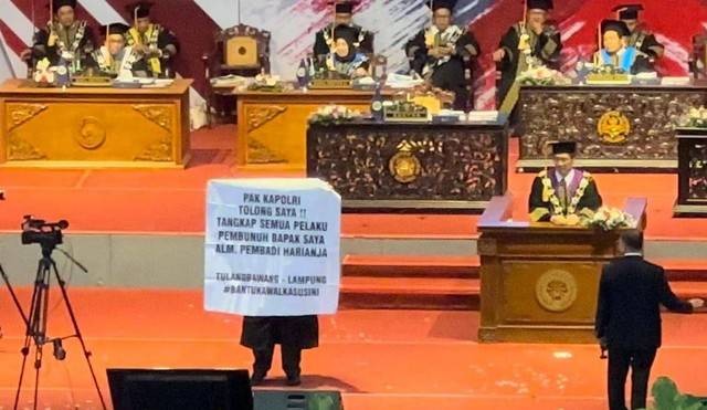 Seorang wisudawan membentangkan spanduk berisi pesan meminta Kapolri tangkap pelaku pembunuh ayahnya. | Foto: istimewa