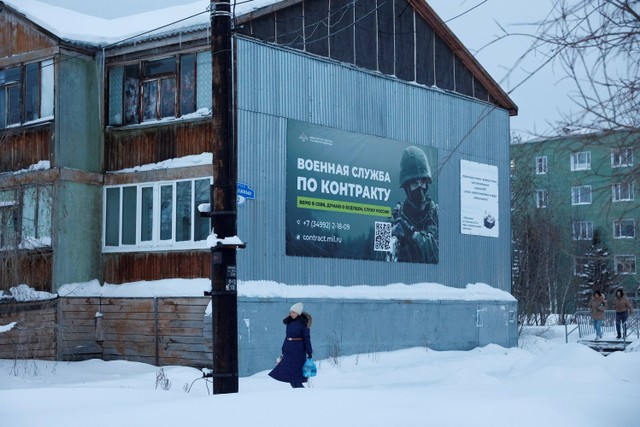 Seorang wanita berjalan melewati spanduk yang mempromosikan dinas militer berdasarkan kontrak dengan Angkatan Bersenjata Rusia di pemukiman Kharp di koloni hukuman IK-3, di Wilayah Yamal-Nenets, Rusia. Foto: STR/REUTERS