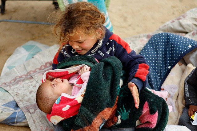 Seorang anak Palestina yang mengungsi yang meninggalkan rumahnya akibat serangan Israel menggendong bayi yang menangis, saat mereka berlindung di tenda kamp di tengah konflik yang sedang berlangsung antara Israel dan Hamas, di Rafah. Foto: Mohammed Salem/Reuters